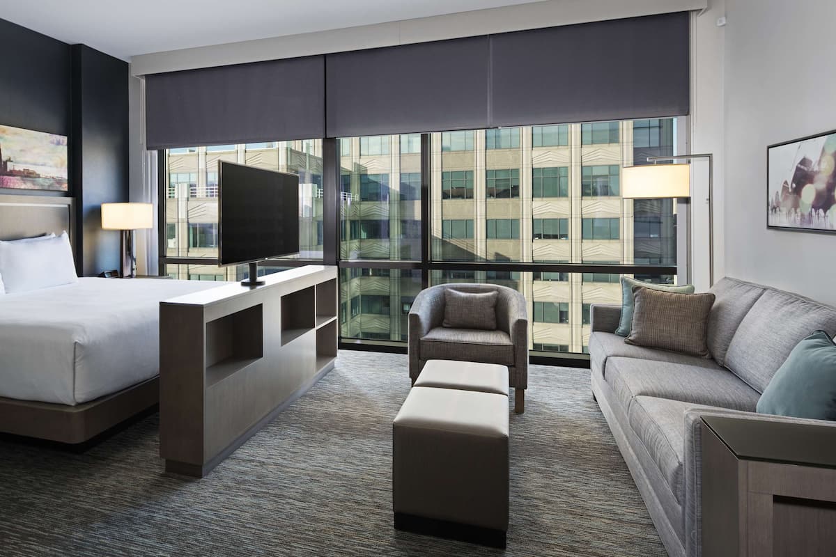 Une chambre d’hôtel avec un grand lit, une télévision, un canapé et un fauteuil. Une grande fenêtre offre une vue sur les bâtiments environnants.