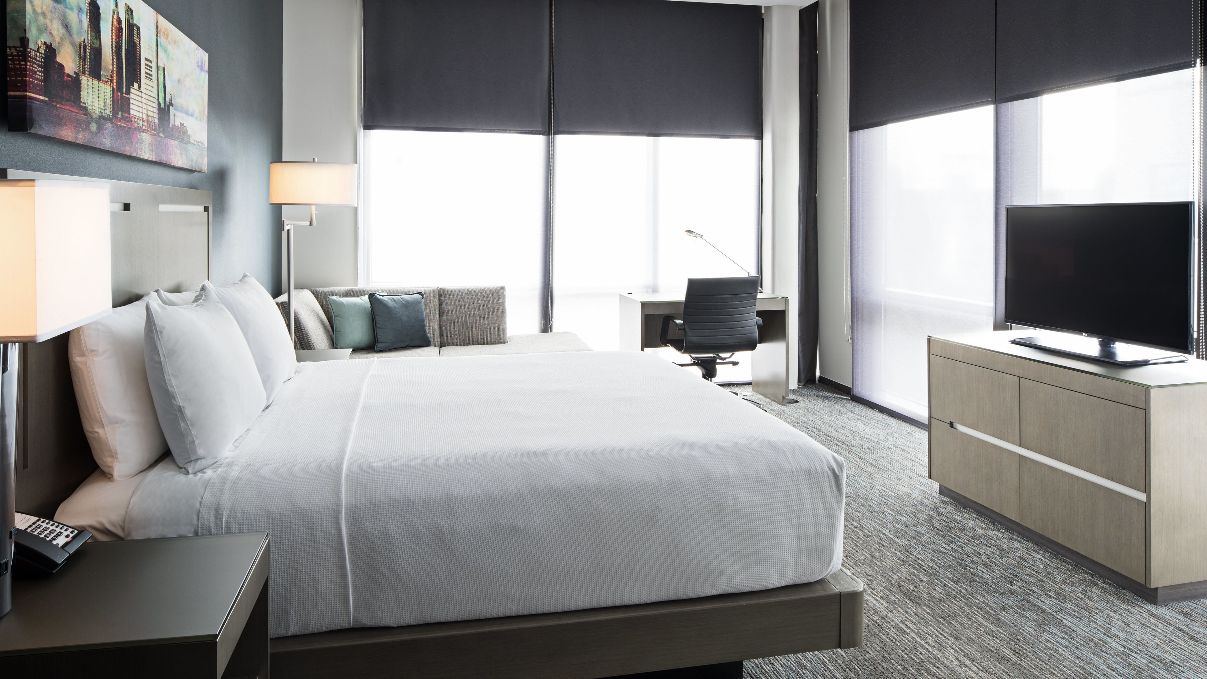 Habitación de hotel con una cama grande, un escritorio con silla, un televisor y una ventana con persianas.