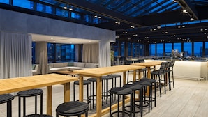 A modern bar area with wooden tables and black bar stools, large windows with curtains, and a city view.