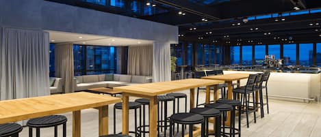 A modern bar area with wooden tables and black bar stools, large windows with curtains, and a city view.