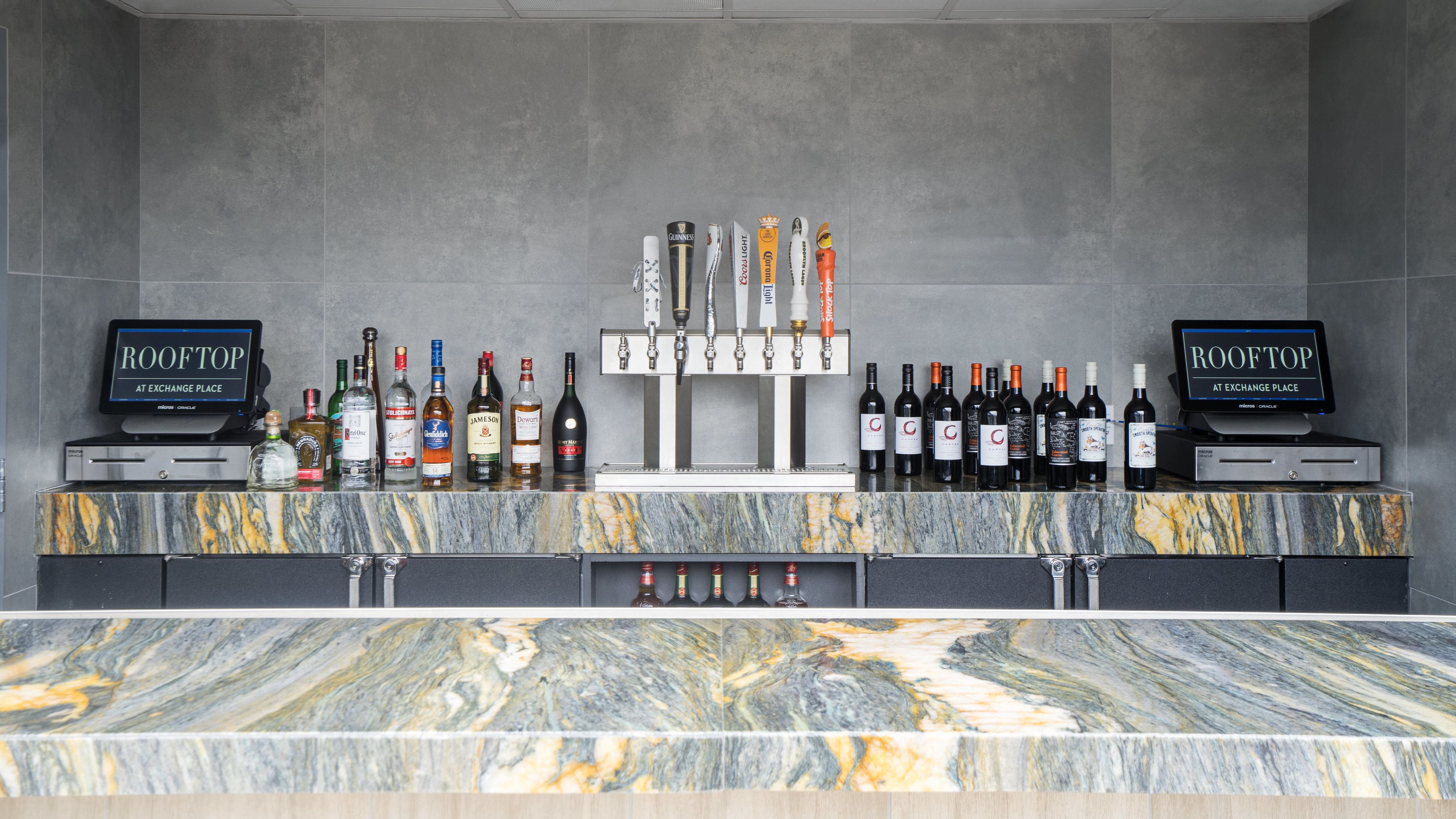 Un espace bar doté d’un comptoir en marbre, de nombreuses bouteilles d’alcool et de deux écrans affichant le texte « ROOFTOP AT EXCHANGE PLACE ».
