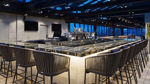 A modern bar area with a marble countertop, black bar stools, and a city view through large windows.