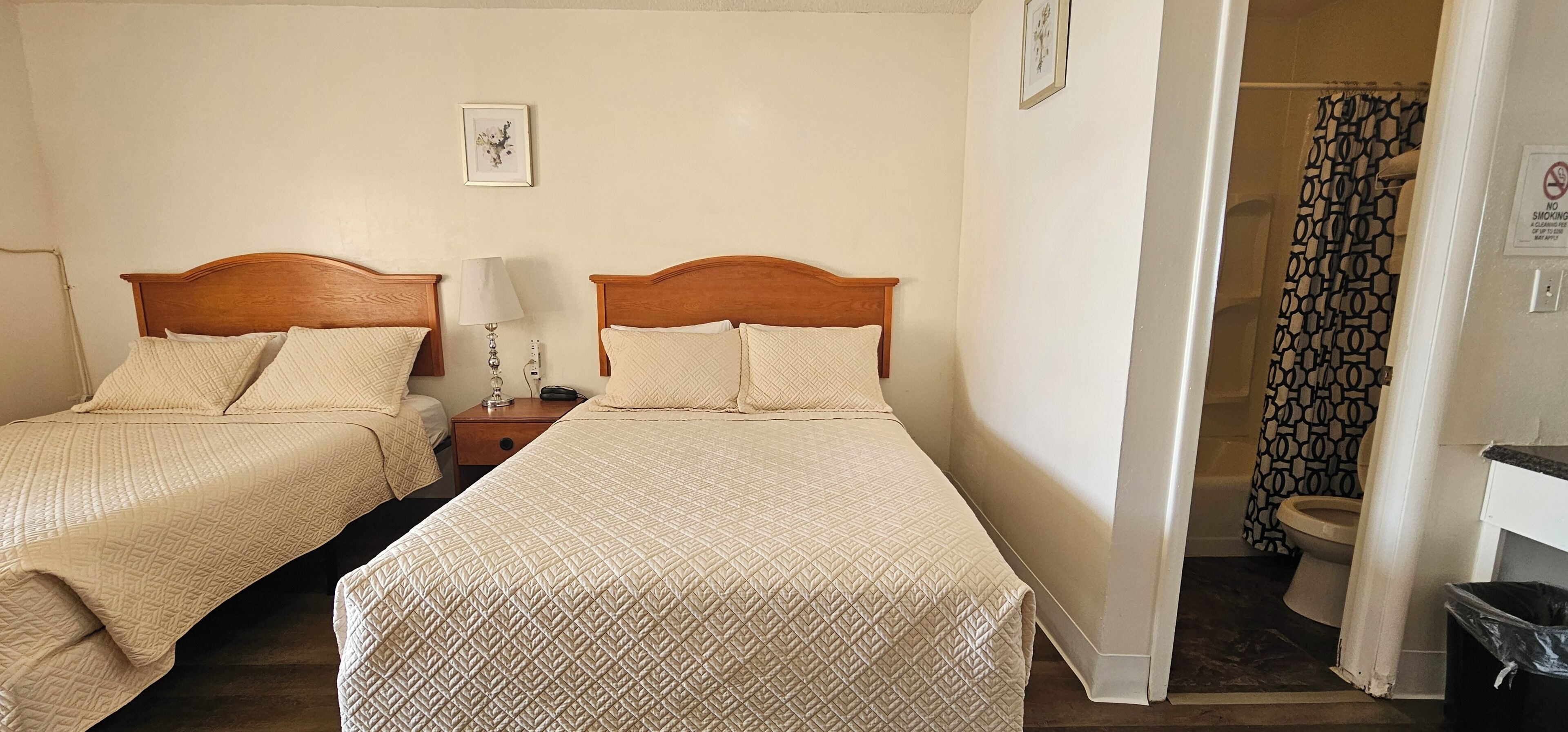 A hotel room with two beds, a nightstand, a lamp, a framed picture on the wall, and a bathroom with a shower curtain.