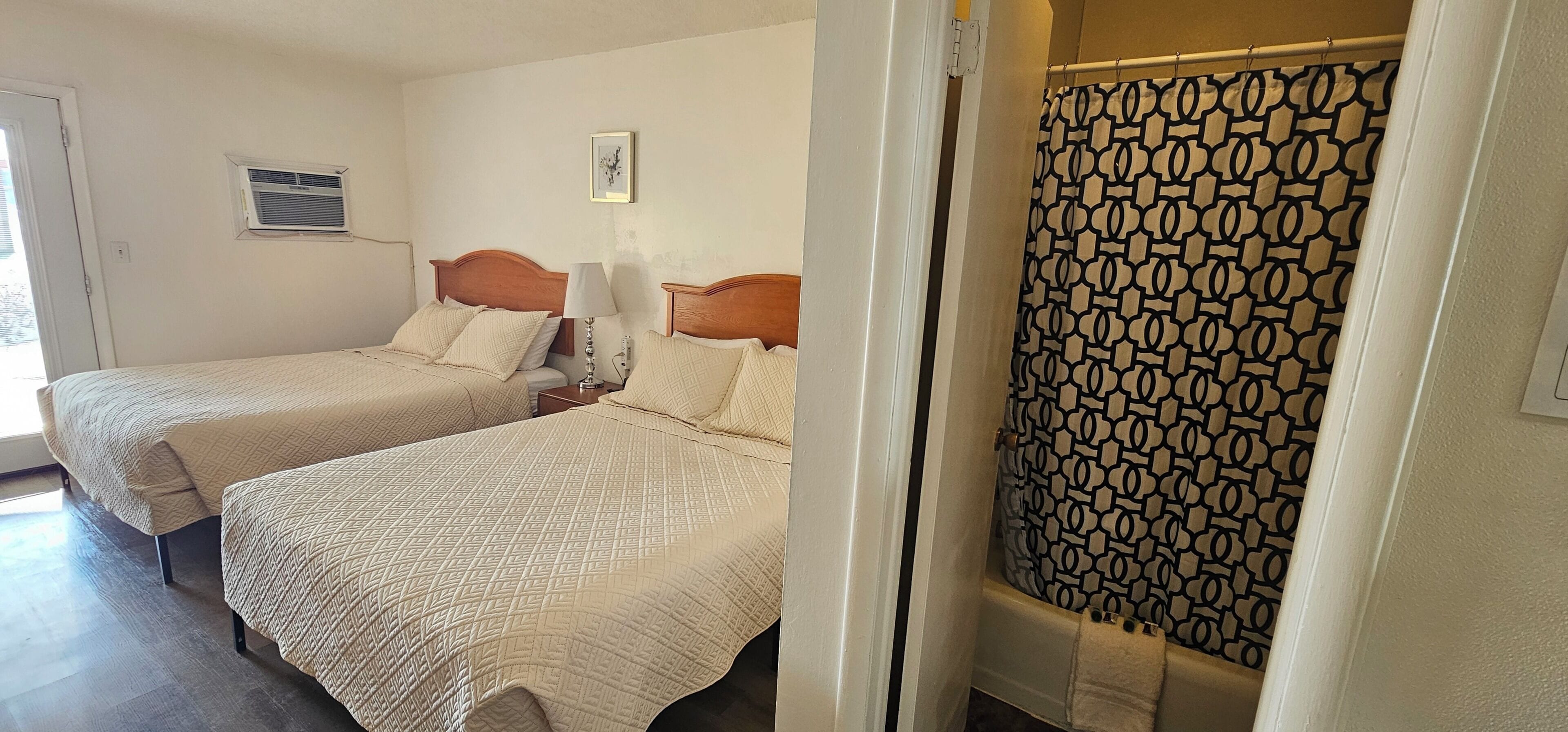 A hotel room with two beds, a shower curtain, and a small window with an air conditioning unit.