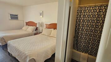 A hotel room with two beds, a shower curtain, and a small window with an air conditioning unit.