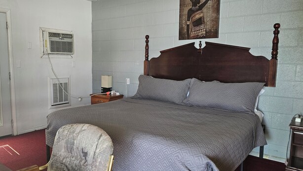 A bedroom with a four-poster bed, a bedside table with a lamp, and a wall-mounted air conditioning unit.