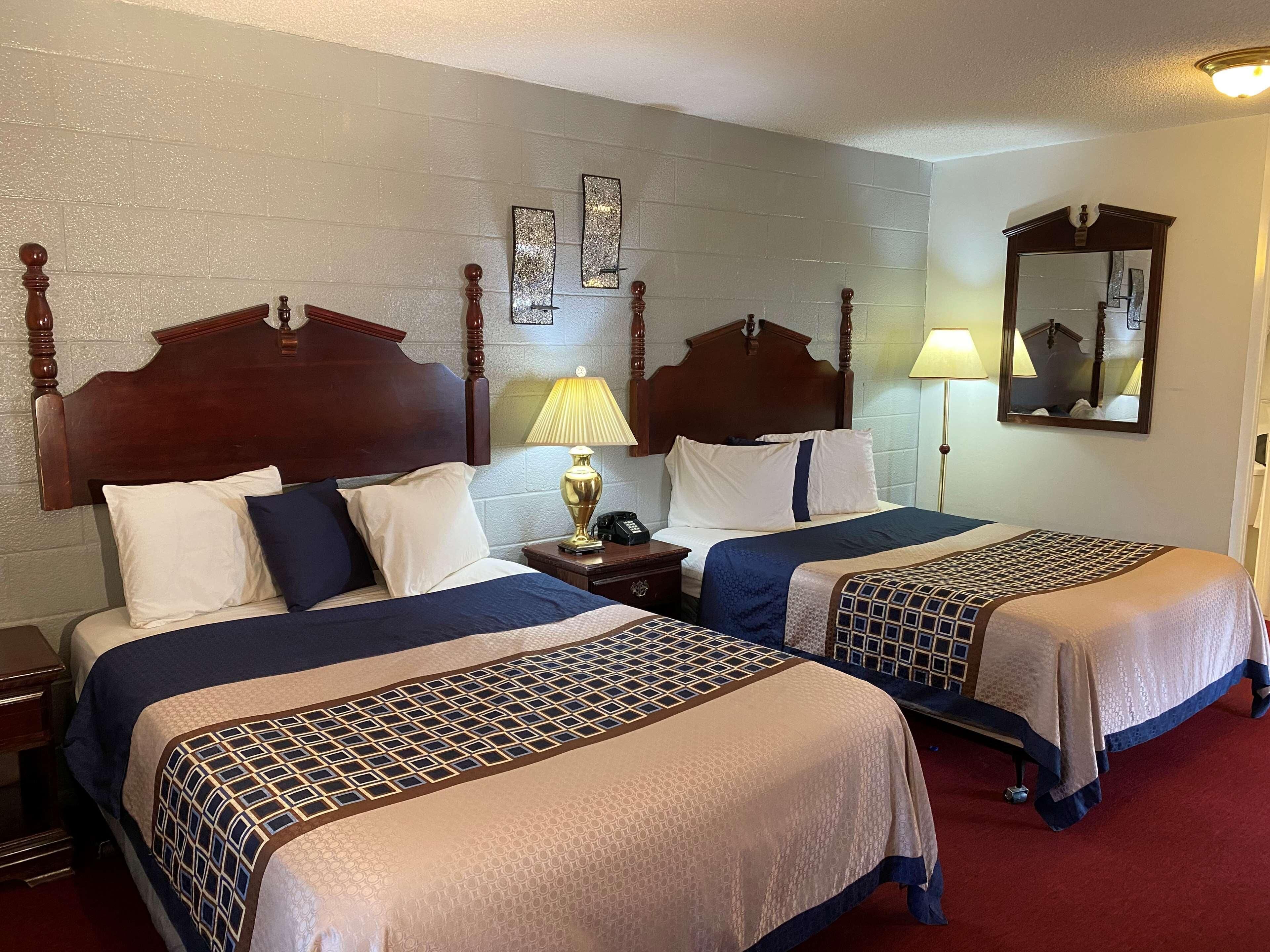 A hotel room with two beds, wooden headboards, a nightstand with a lamp, a mirror, and a wall clock.