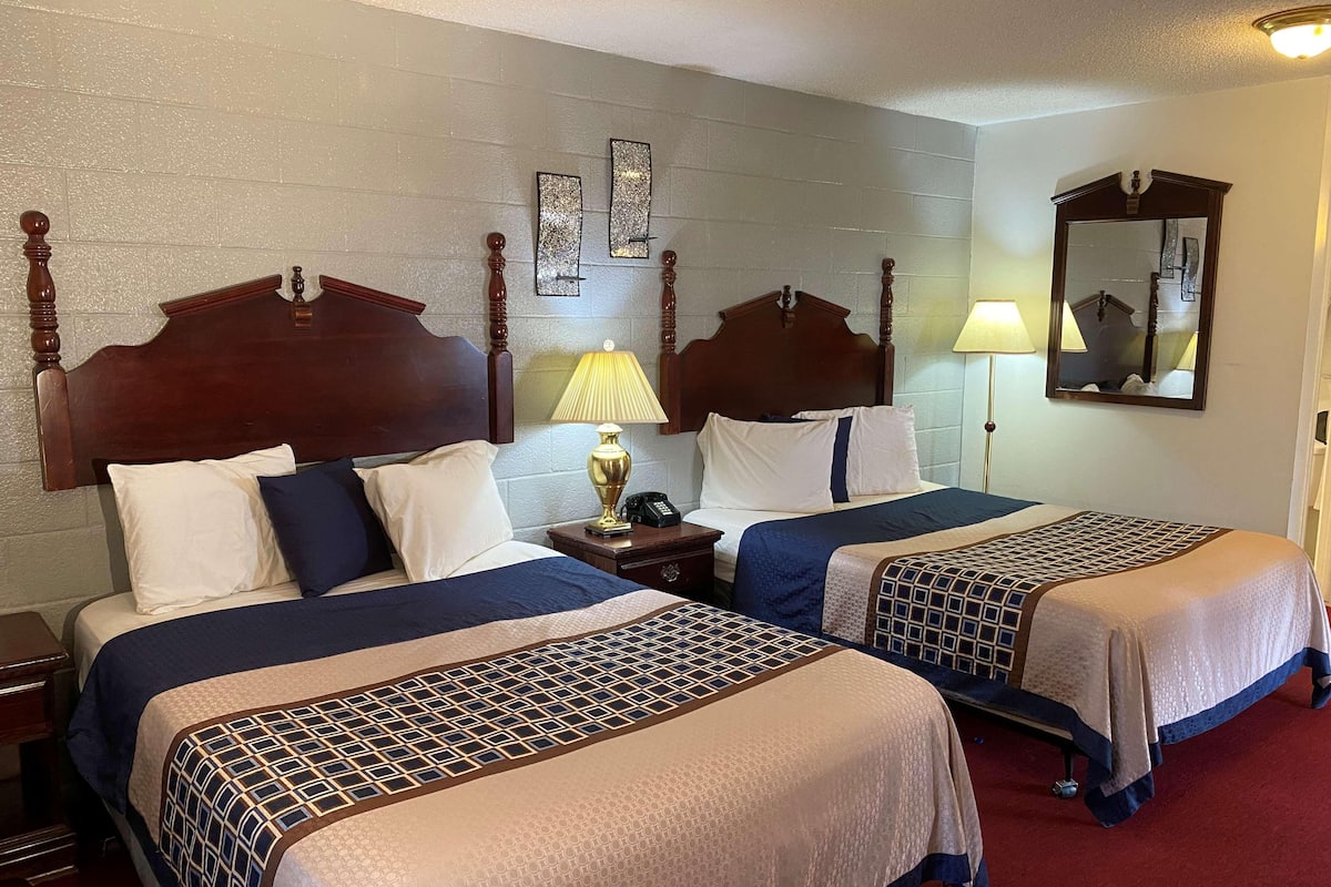 A hotel room with two beds, wooden headboards, a nightstand with a lamp, a mirror, and a wall clock.