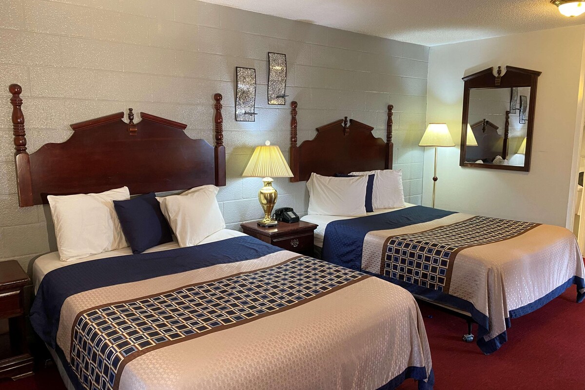 A hotel room with two beds, wooden headboards, a nightstand with a lamp, a mirror, and a wall clock.