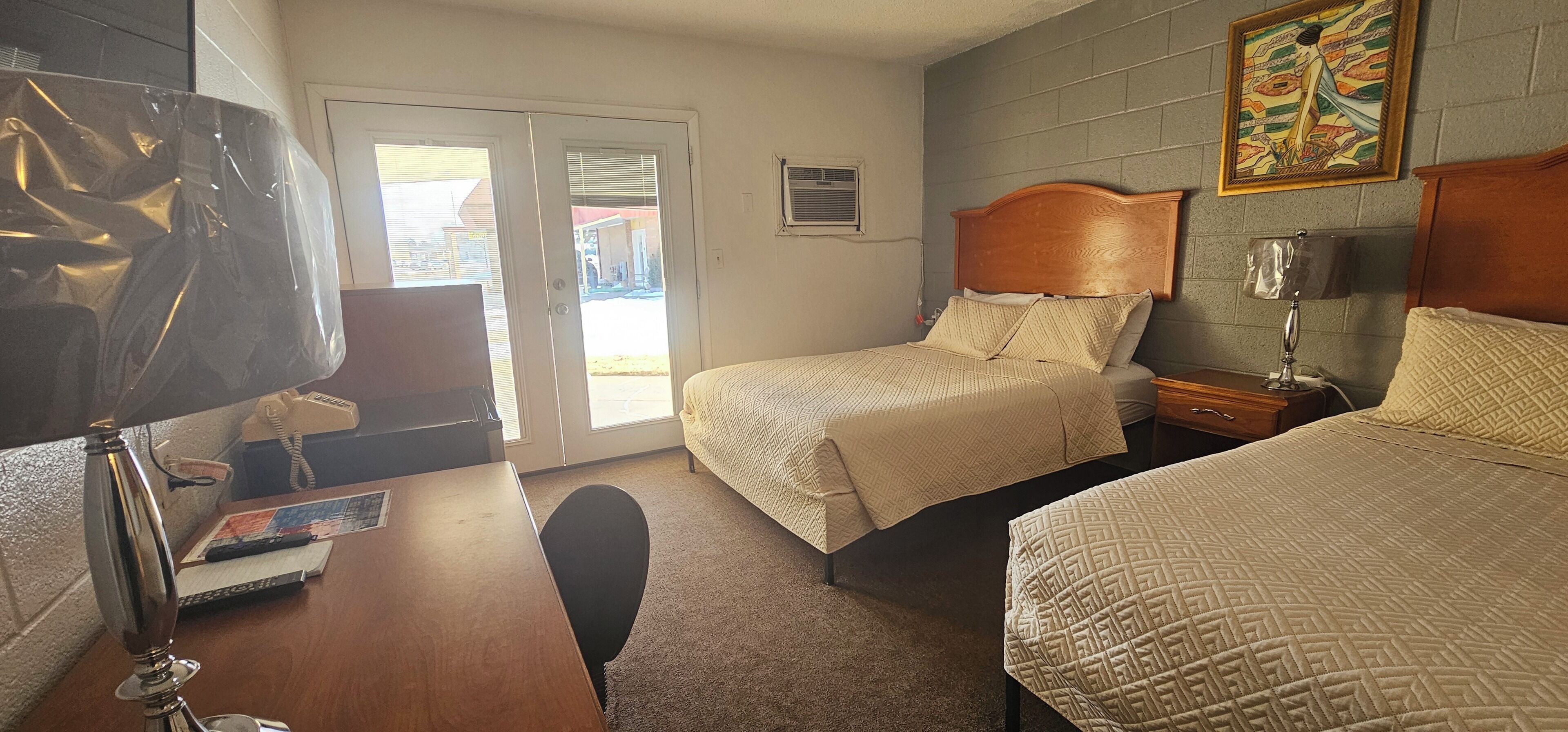 A hotel room with two beds, a desk with a lamp, a chair, and a window with blinds.