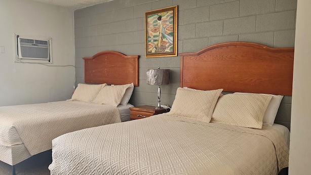 A room with two beds, a wall-mounted air conditioning unit, a framed picture, and a bedside lamp.