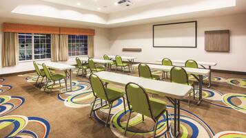 Un salón de conferencias con varias mesas y sillas, una pantalla de proyector y un amplio ventanal con cortinas.