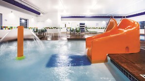 Un tobogán acuático interior con un tubo transparente y un tobogán naranja, rodeado de una zona de piscina con suelo de baldosas y algunas sillas.