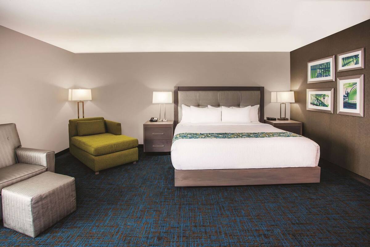 A hotel room with a large bed, two bedside tables, a green armchair, a grey armchair, and a patterned carpet.