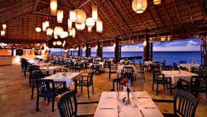 Een ruime restaurantzaal met gedekte tafels, een rieten dak en uitzicht op de oceaan.