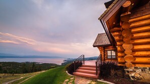 Uma cabana de toras com um caminho de pedra que leva a um lance de escadas, com vista para um corpo d'água e colinas.