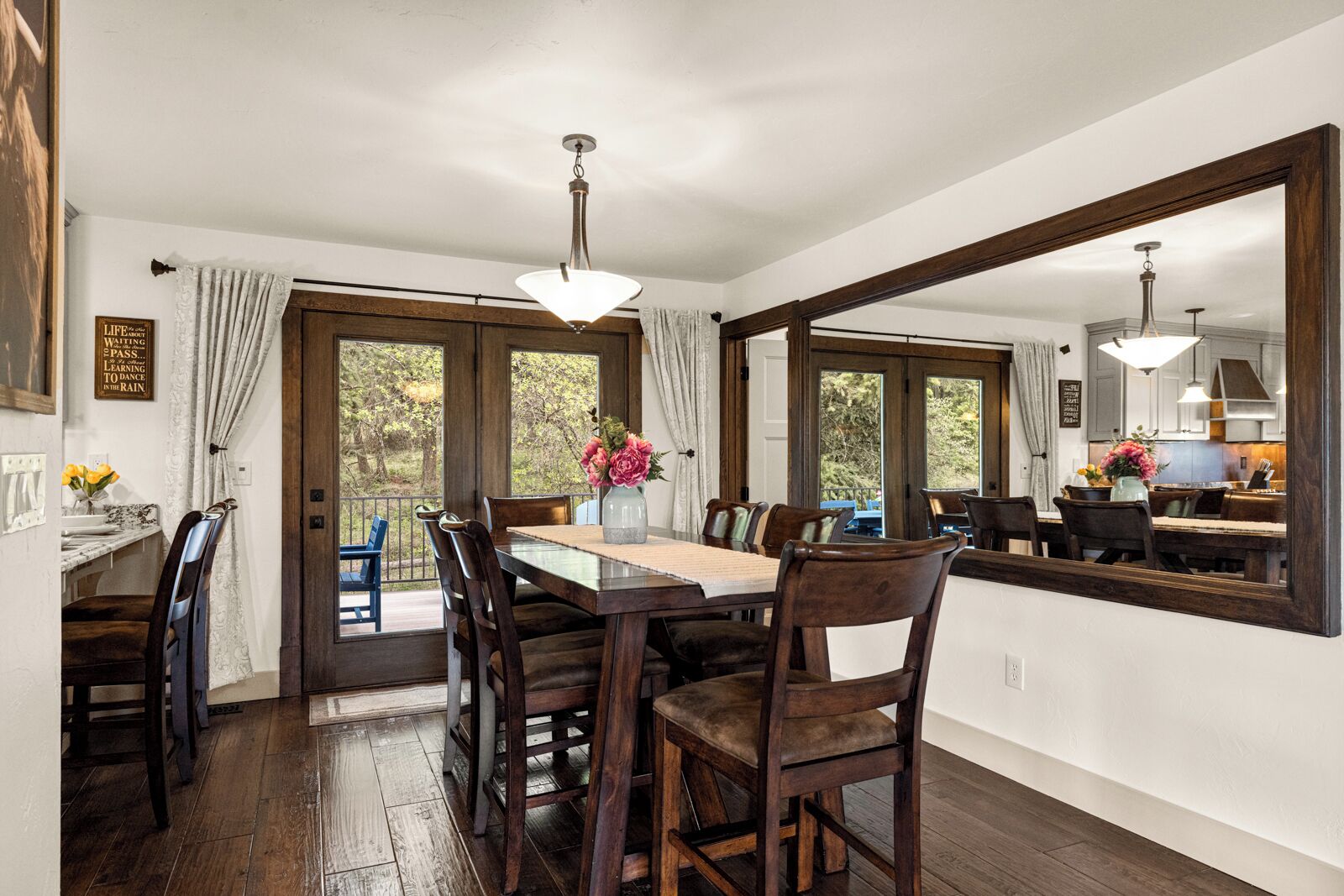 A dining area with a wooden table, chairs, and a vase of flowers. There is a large mirror on the wall and a view of the outdoors through the windows.