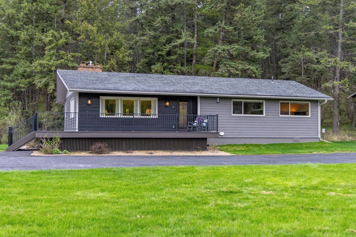 A modern two-story house with a covered porch and a small deck, surrounded by a grassy yard and dense trees.