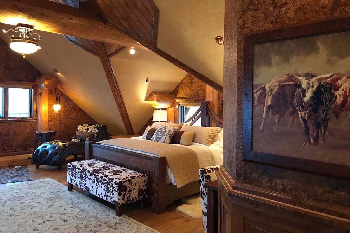 A bedroom with a sloped ceiling, wooden beams, a bed with a cow-patterned ottoman, a painting of cattle, and a large window.