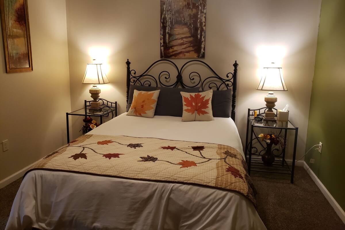 Un lit bien fait, recouvert d’une couvre-lit à motifs de feuilles, deux lampes de chevet, un tableau encadré au-dessus du lit et une petite table avec un vase de fleurs à côté du lit.