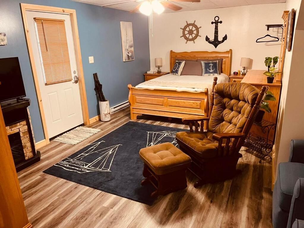 Un dormitorio con una cama de madera, un sillón tapizado, un pequeño reposapiés, un aparador de madera, un televisor y una chimenea.