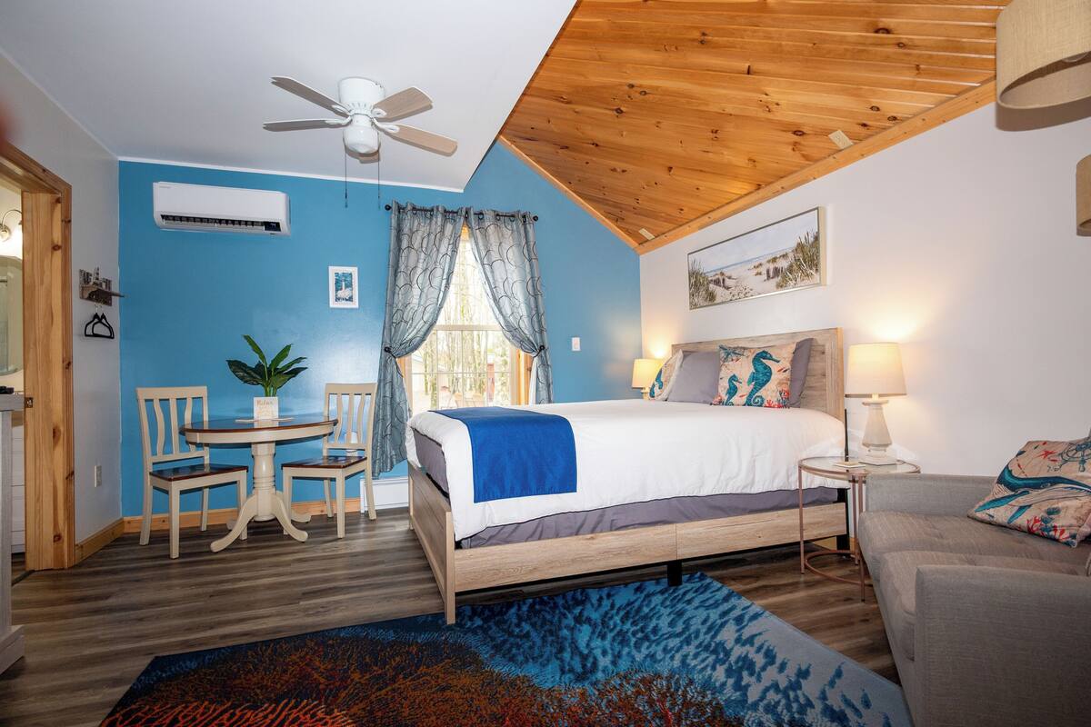 Une chambre à coucher avec un lit, un ventilateur de plafond, une table avec des chaises, une plante et une fenêtre avec des rideaux.