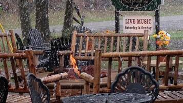 A rustic outdoor seating area with wooden benches and metal chairs, a fire pit, and a sign indicating live music on Saturday and Sunday.