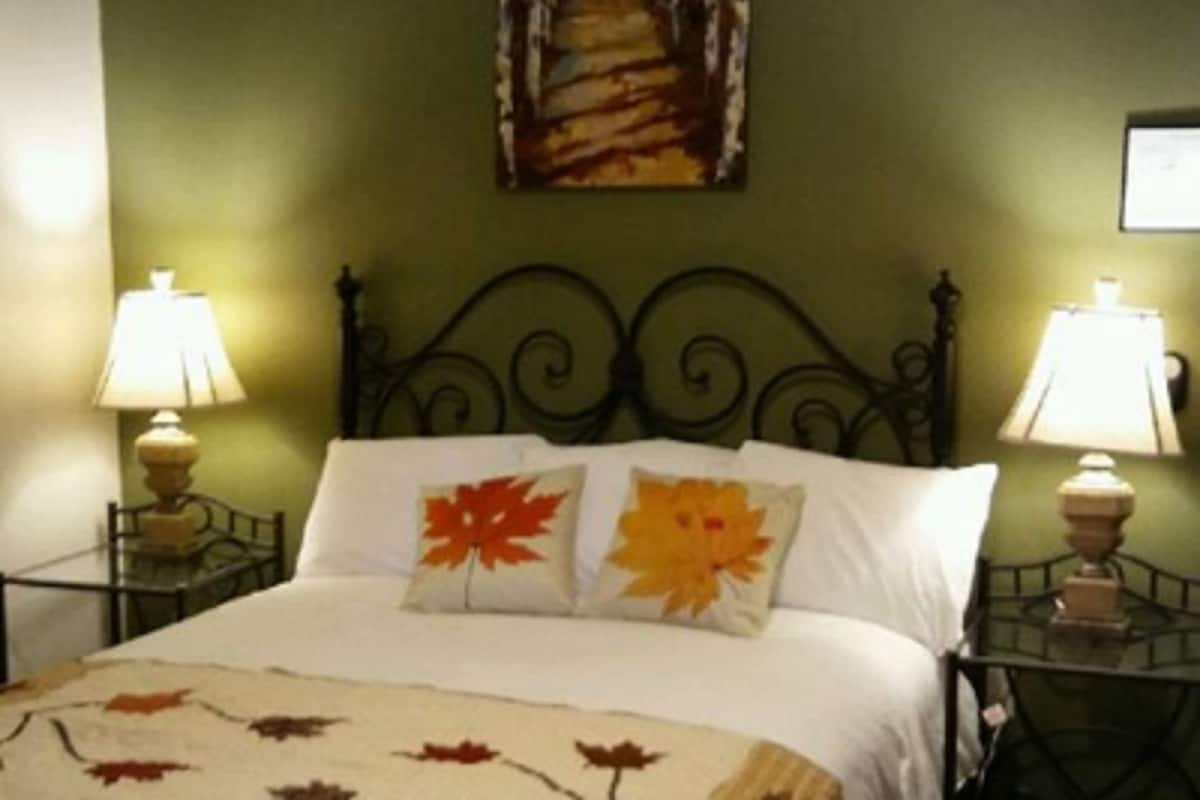 Un lit avec une literie blanche et deux oreillers, dont l’un est orné d’un motif représentant une feuille d’érable. Il y a deux tables de chevet avec des lampes, et un tableau représentant un sentier forestier est accroché au mur.