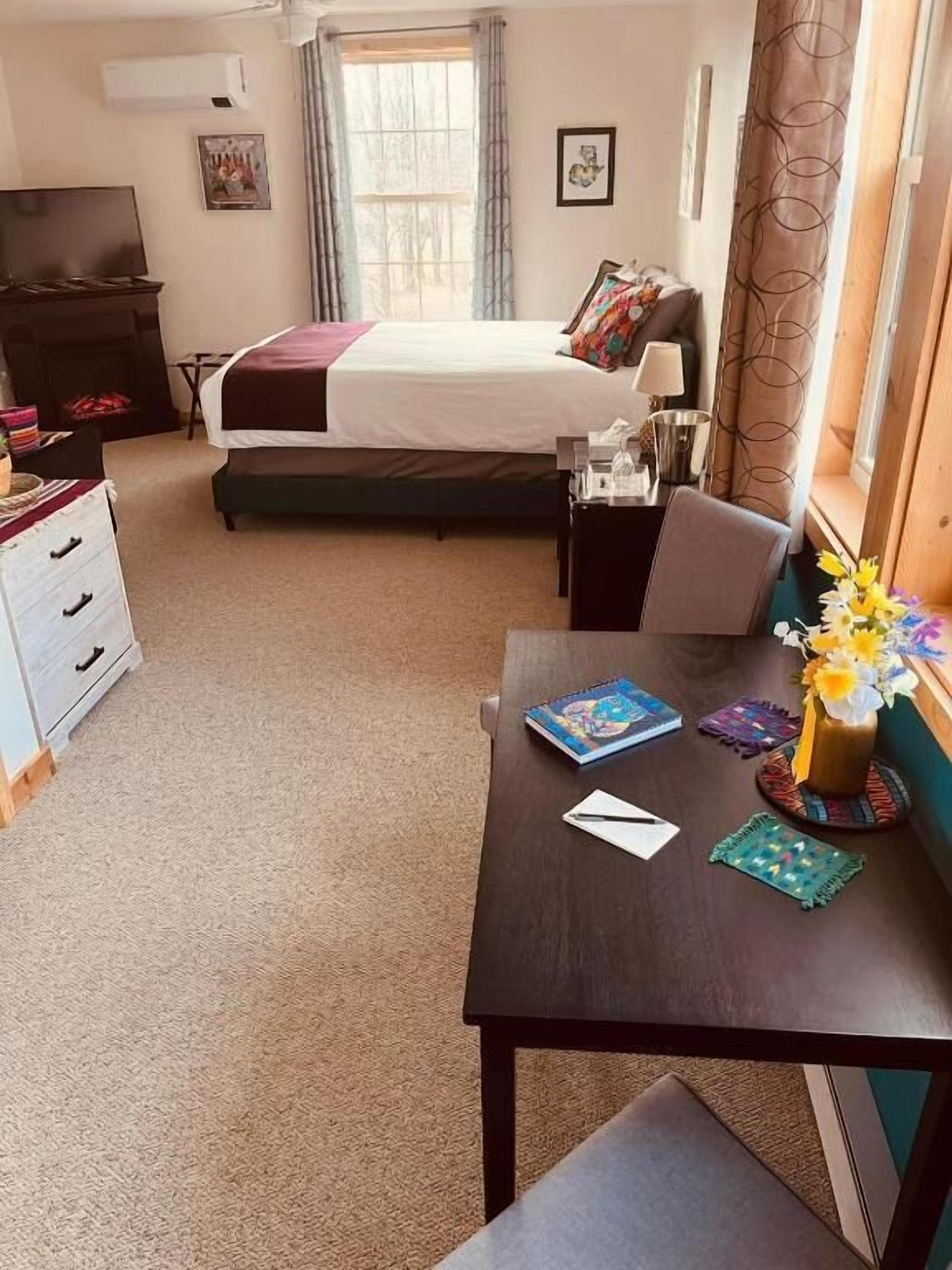Quarto de hotel com cama, escrivaninha, cadeira, cômoda, televisão e janela com cortinas.