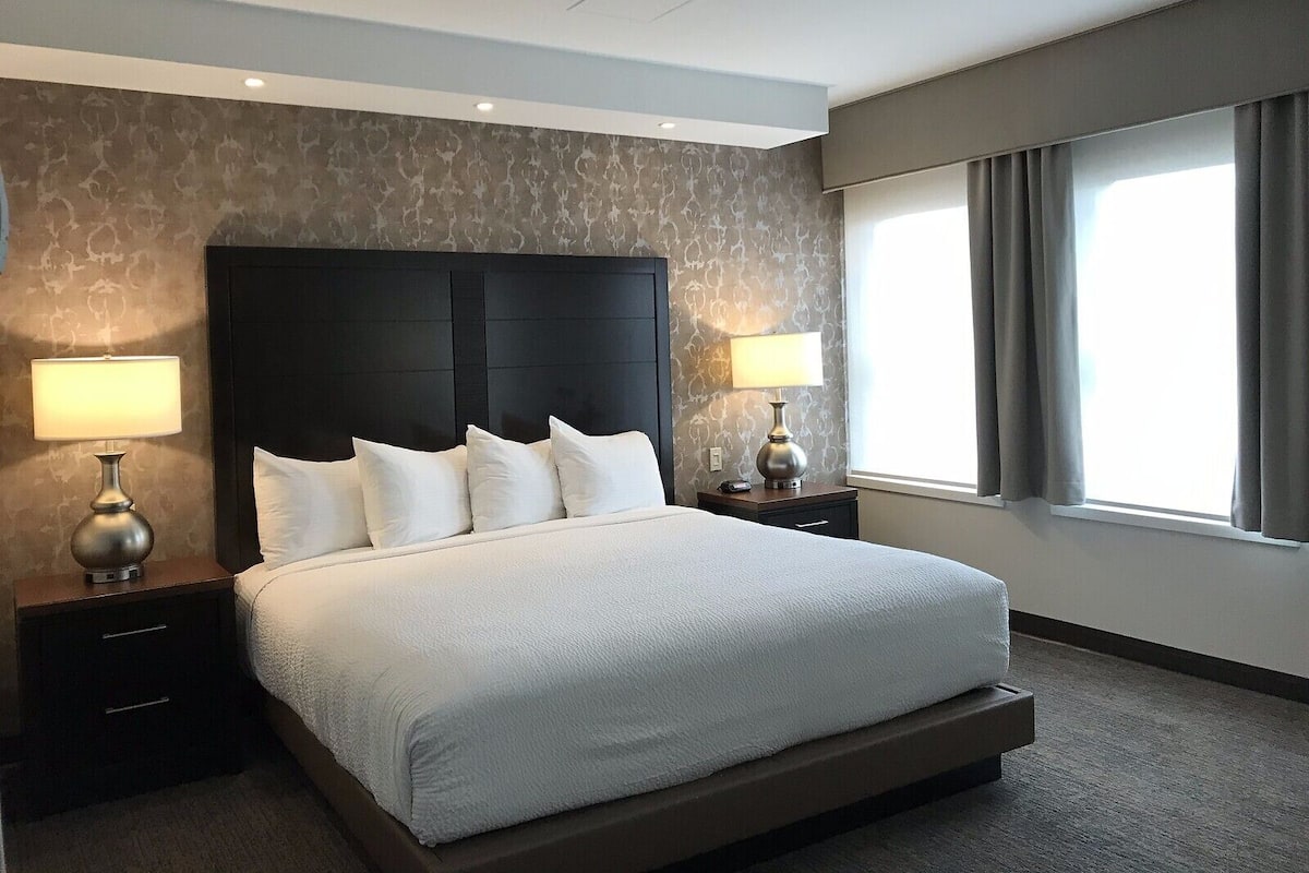 Habitación de hotel con una cama grande, dos mesitas de noche con lámparas, un cabecero y una ventana con cortinas.
