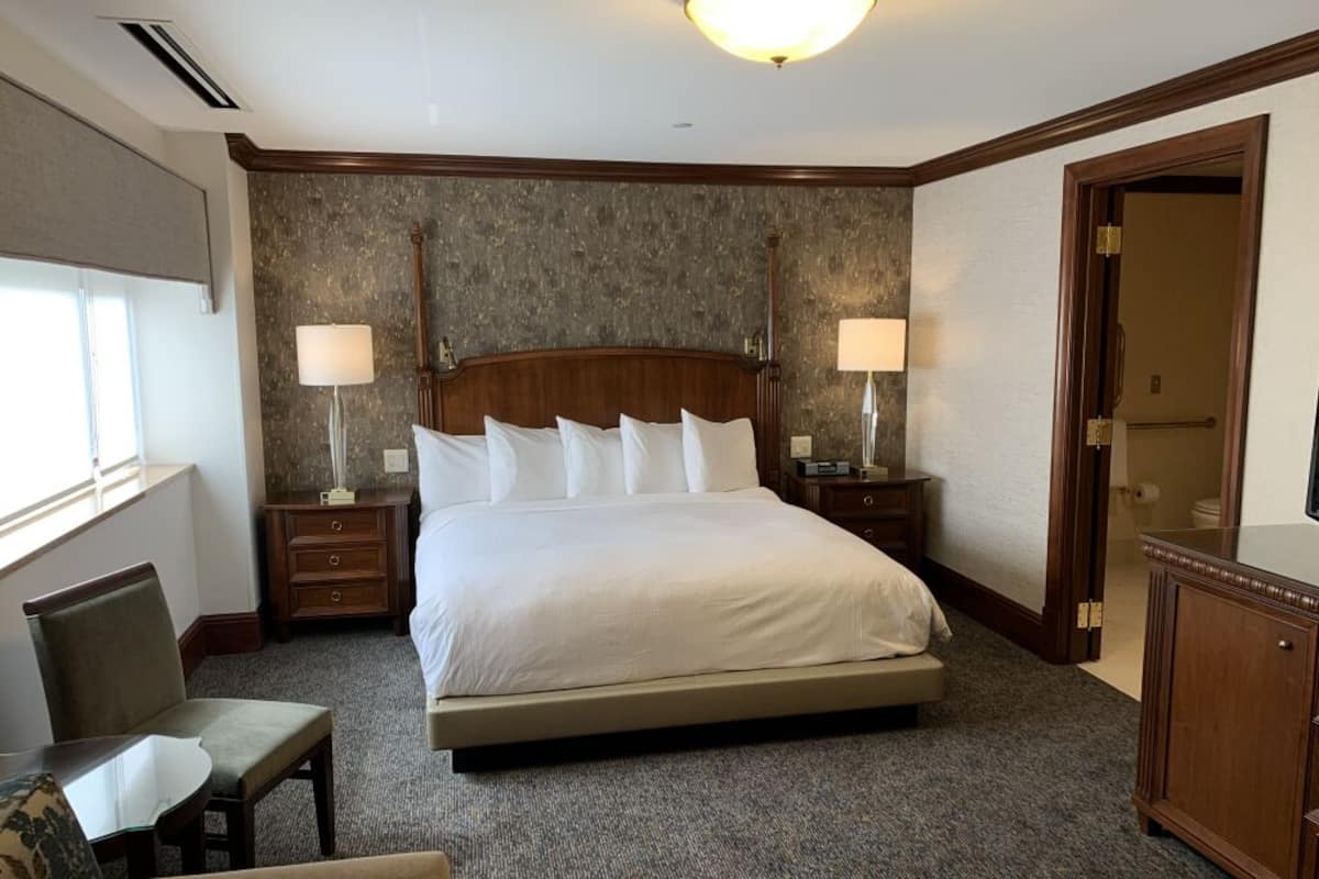 Una habitación de hotel con una cama grande, dos mesitas de noche, una silla y un aparador.