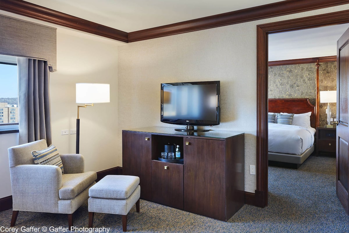 Una habitación de hotel con televisión sobre un mueble de madera, una cama con almohadas, una silla con cojín y una mesita de noche con lámpara.