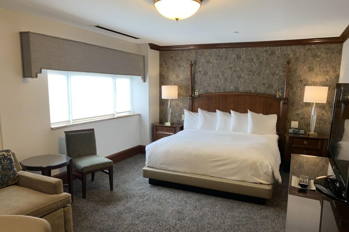 Habitación de hotel con una cama grande, dos mesitas de noche con lámparas, una silla, un escritorio pequeño con televisor y una ventana con persianas.