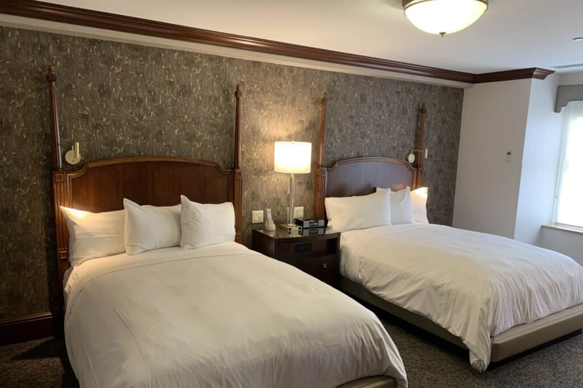 Habitación de hotel con dos camas, cabeceros de madera, una mesita de noche con lámpara y pared con textura.