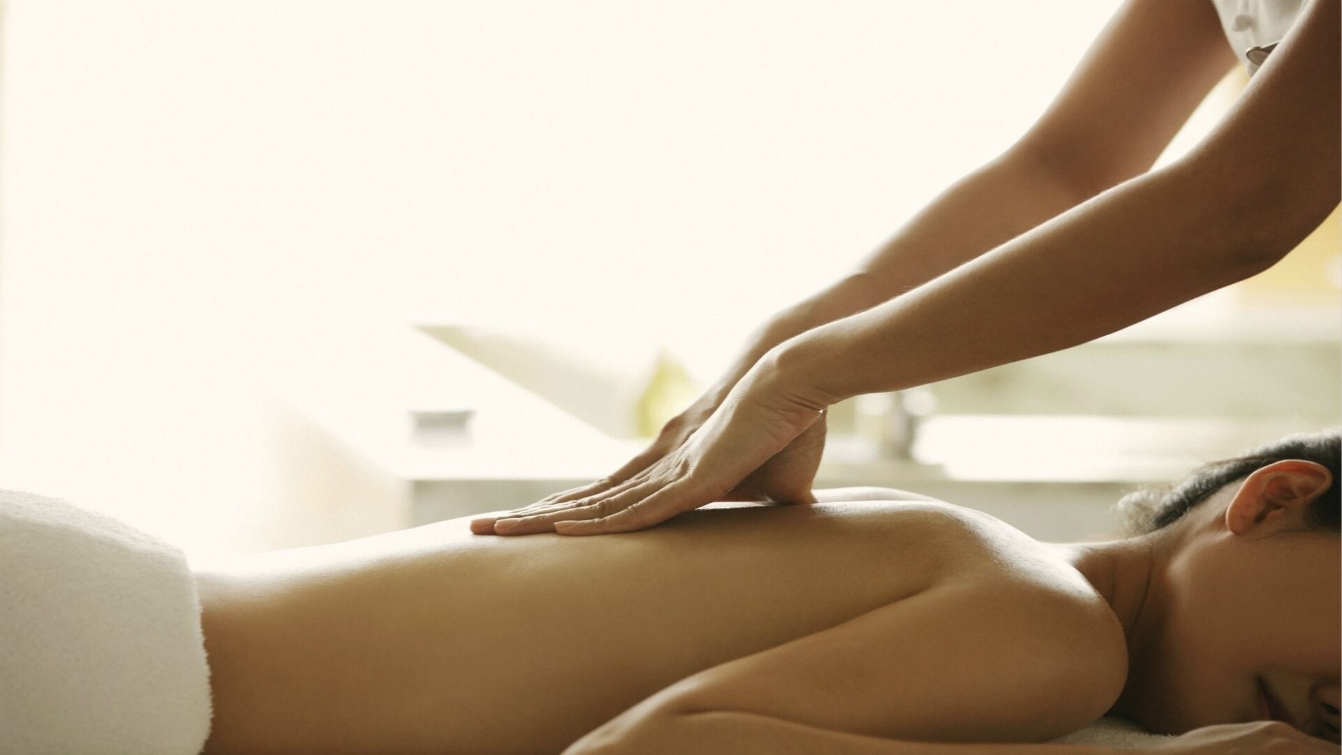 Eine Person erhält eine Rückenmassage in einem Raum, im Hintergrund ein weißes Bett und ein kleiner Tisch.