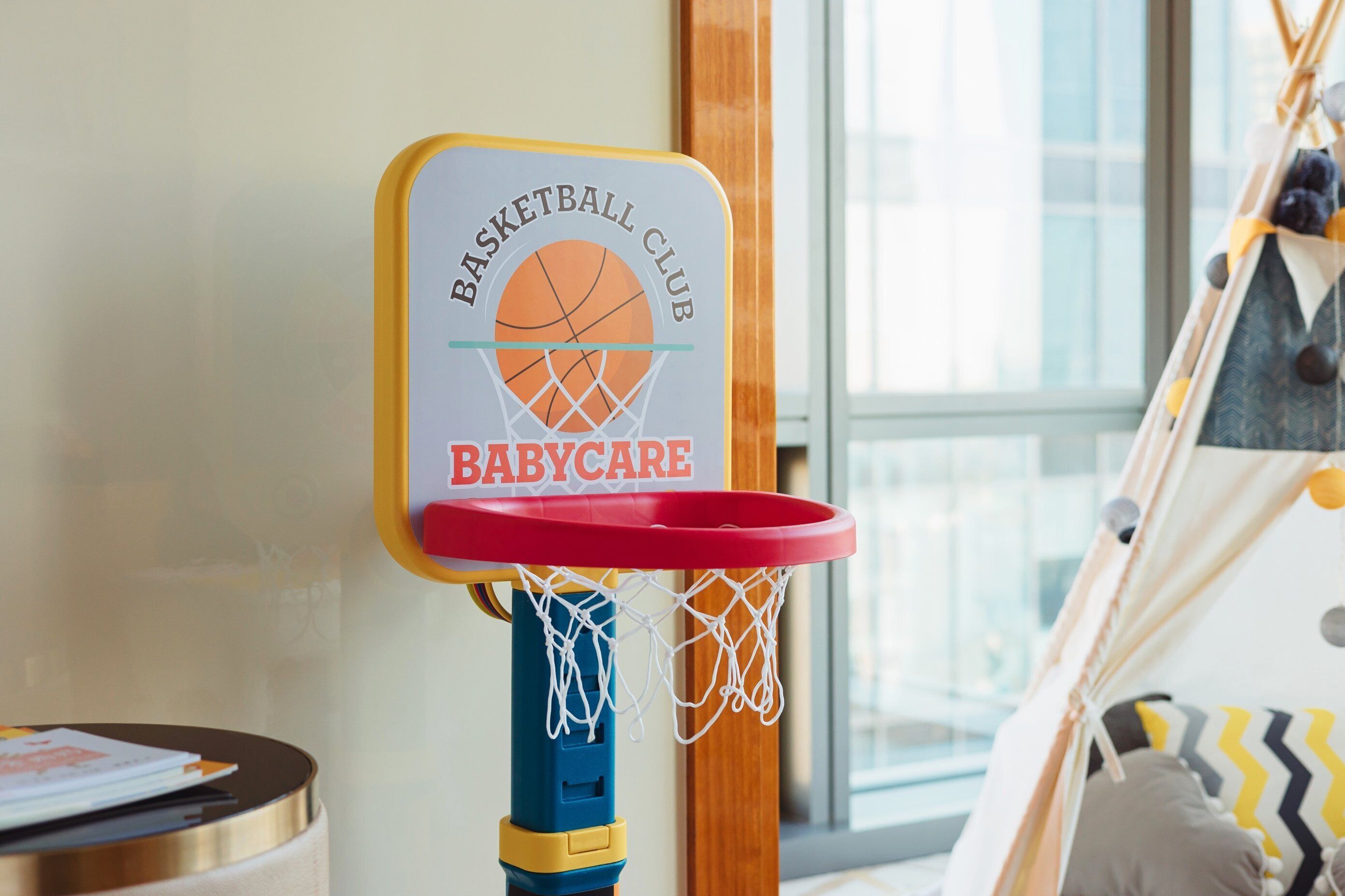 A basketball hoop with a sign reading "Basketball Club Babycare" in a room with a window and a tent-like structure in the background.