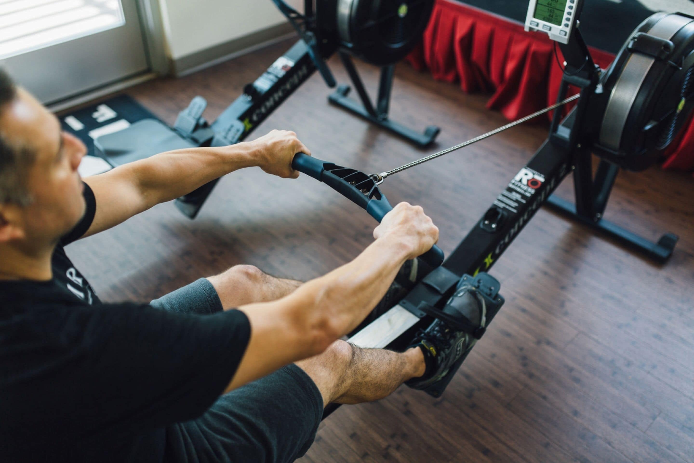 A person using a rowing machine in a gym with a weight stack and a red towel in the background.