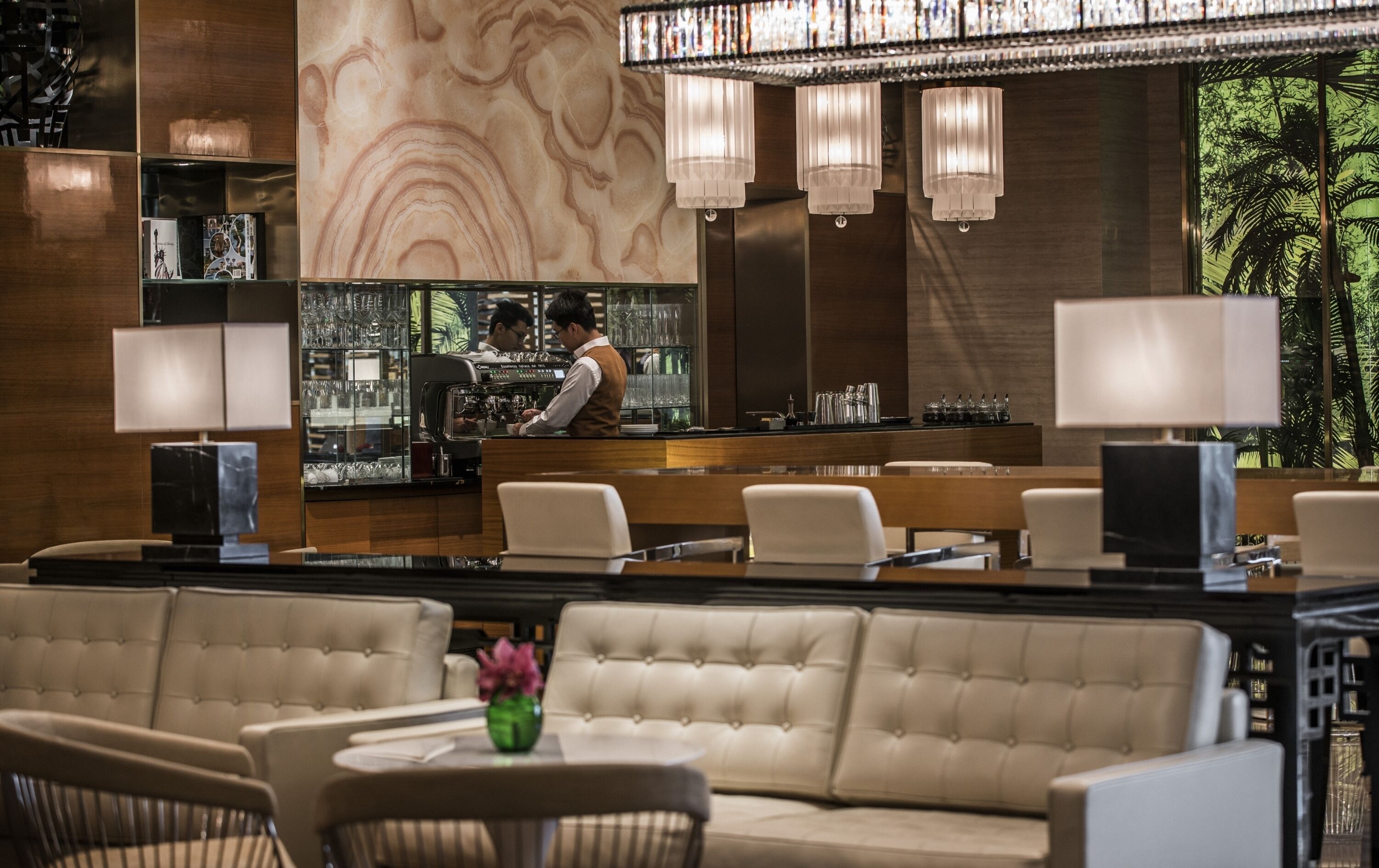 A modern bar area with a bartender, cushioned seating, and a large window with a view of palm trees.