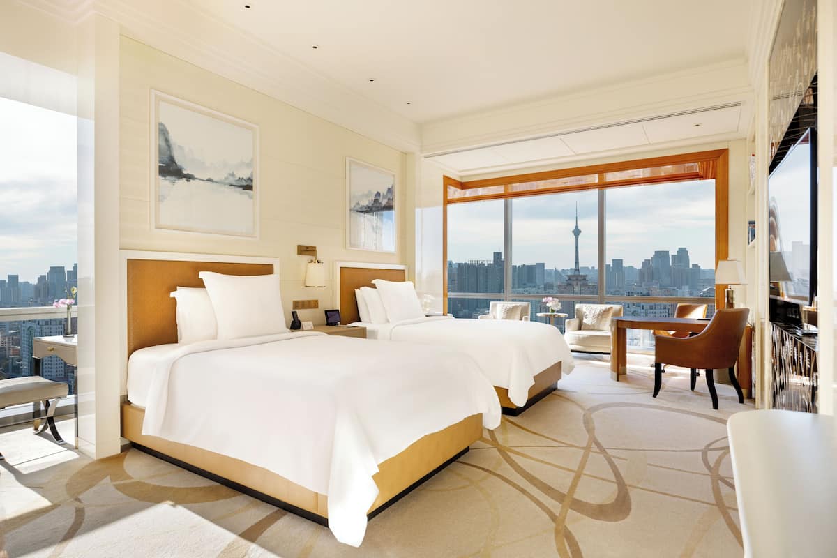 Amplia habitación de hotel con dos camas grandes, zona de comedor y un ventanal con vistas a la ciudad.