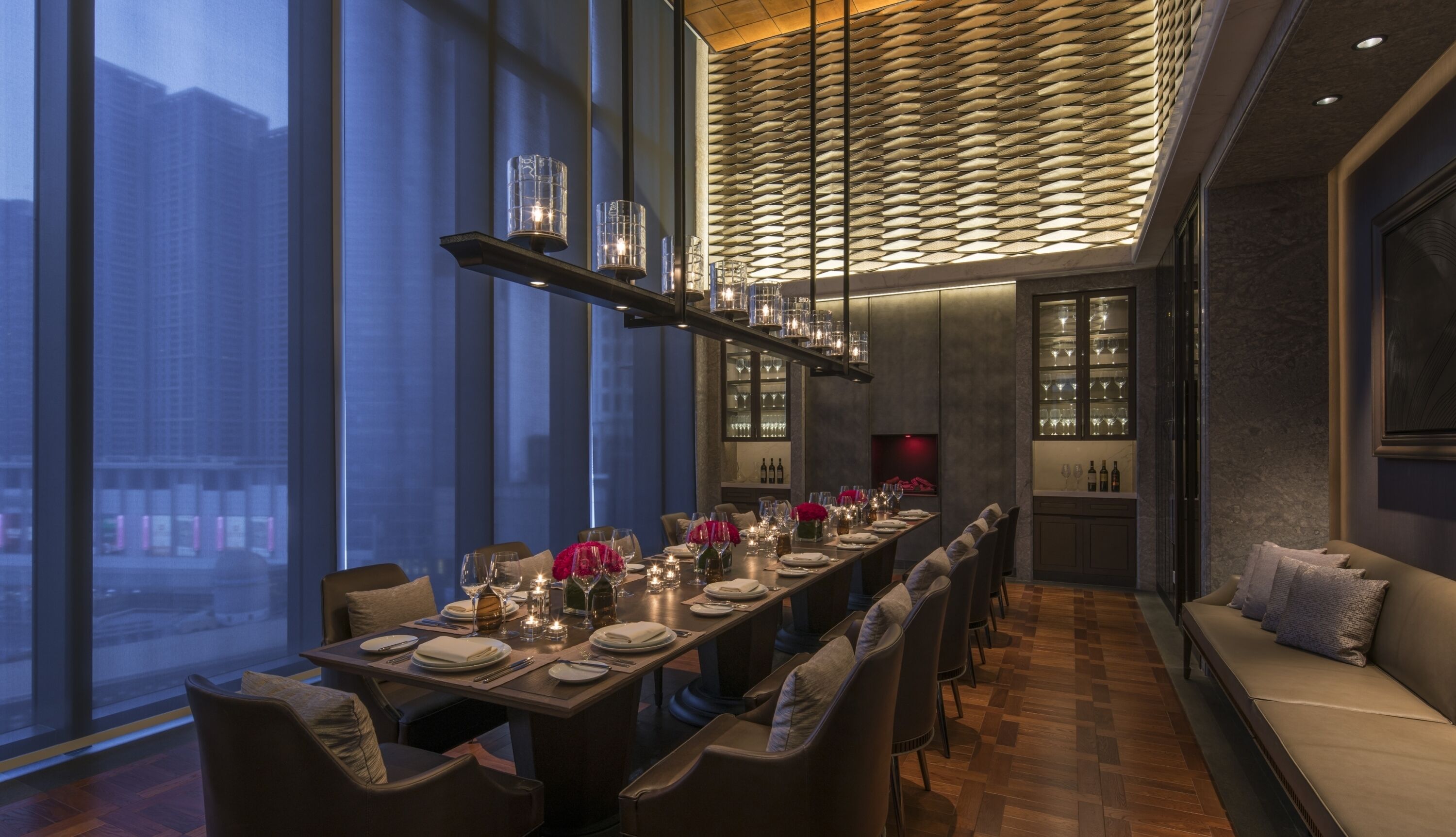 Un comedor moderno con una larga mesa dispuesta para una comida, una zona de estar con bancos acolchados y vistas de la ciudad a través de grandes ventanales.