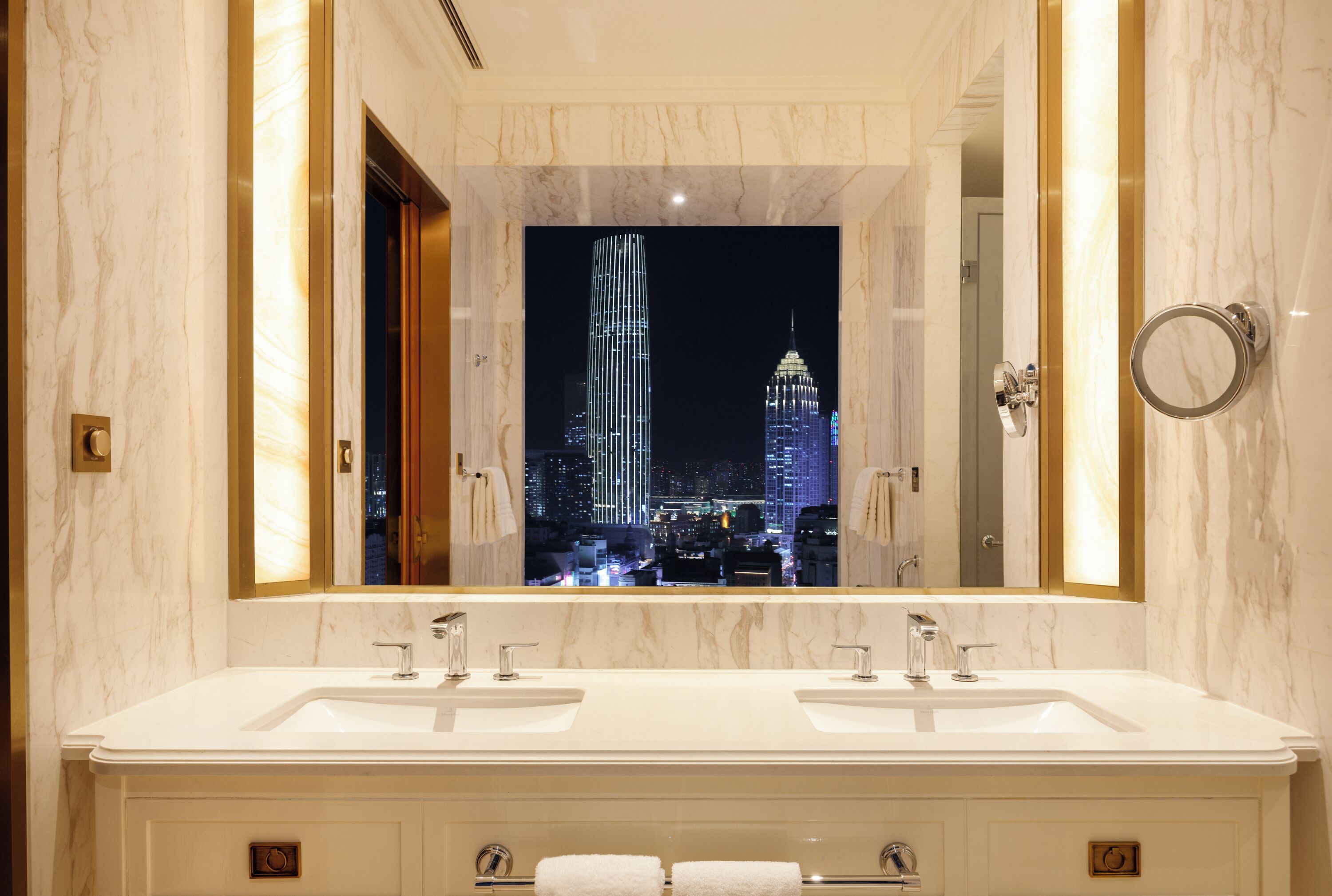 A modern bathroom with two sinks, a large mirror, and a view of a cityscape at night.