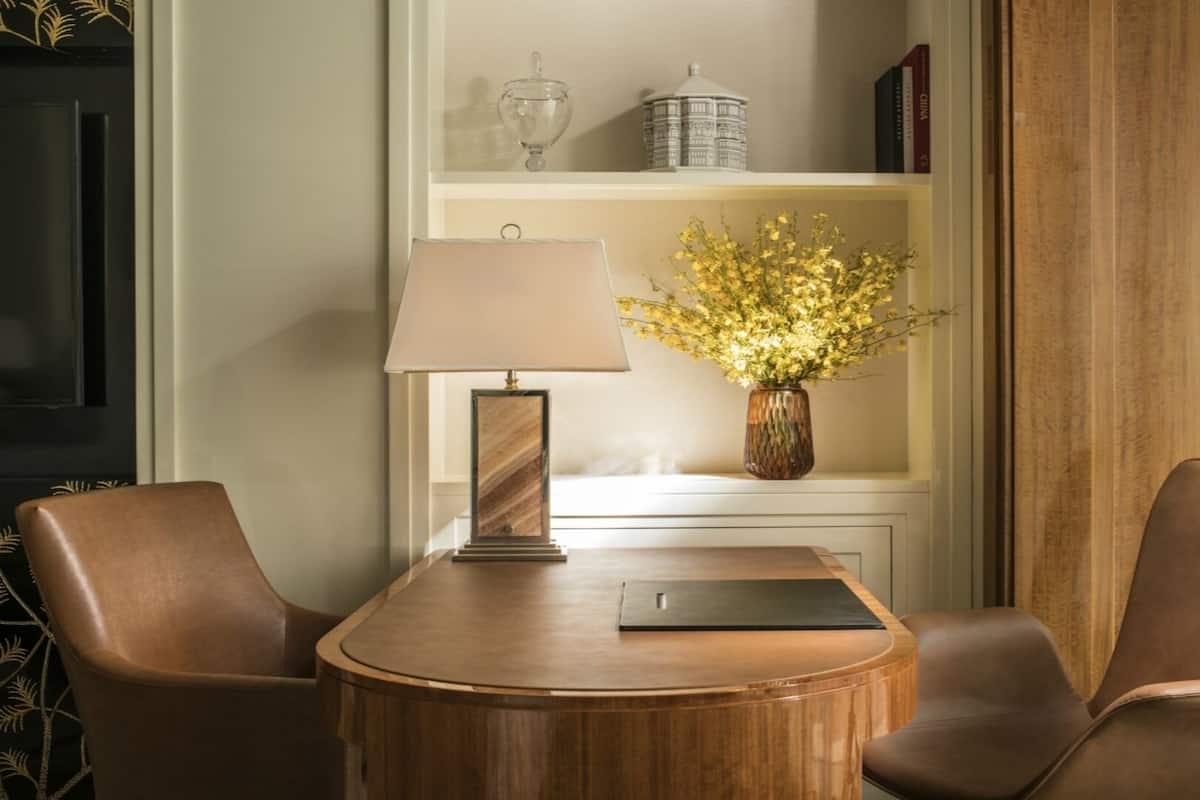 Una mesa redonda de madera con dos sillas de cuero, una lámpara y un florero con flores amarillas.
