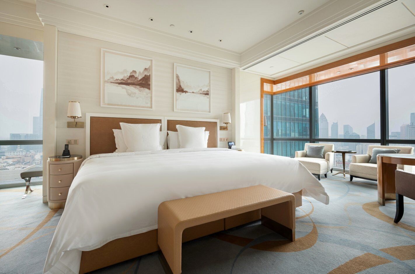 A modern hotel room with a large bed, a bench, a desk, and a view of the cityscape through large windows.
