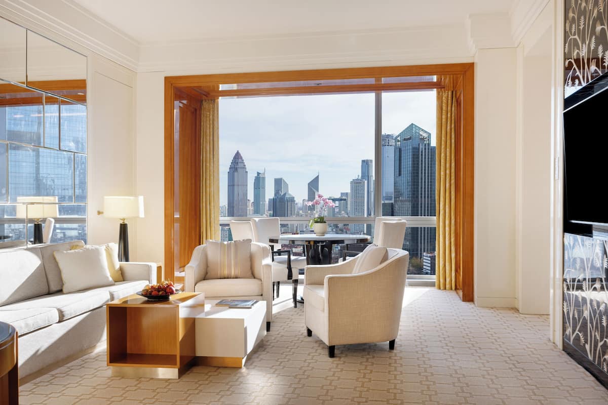 Amplia habitación de hotel con un gran ventanal que ofrece vistas a un horizonte urbano, una zona de estar cómoda con sofá y sillones, y un televisor de pantalla plana en la pared.