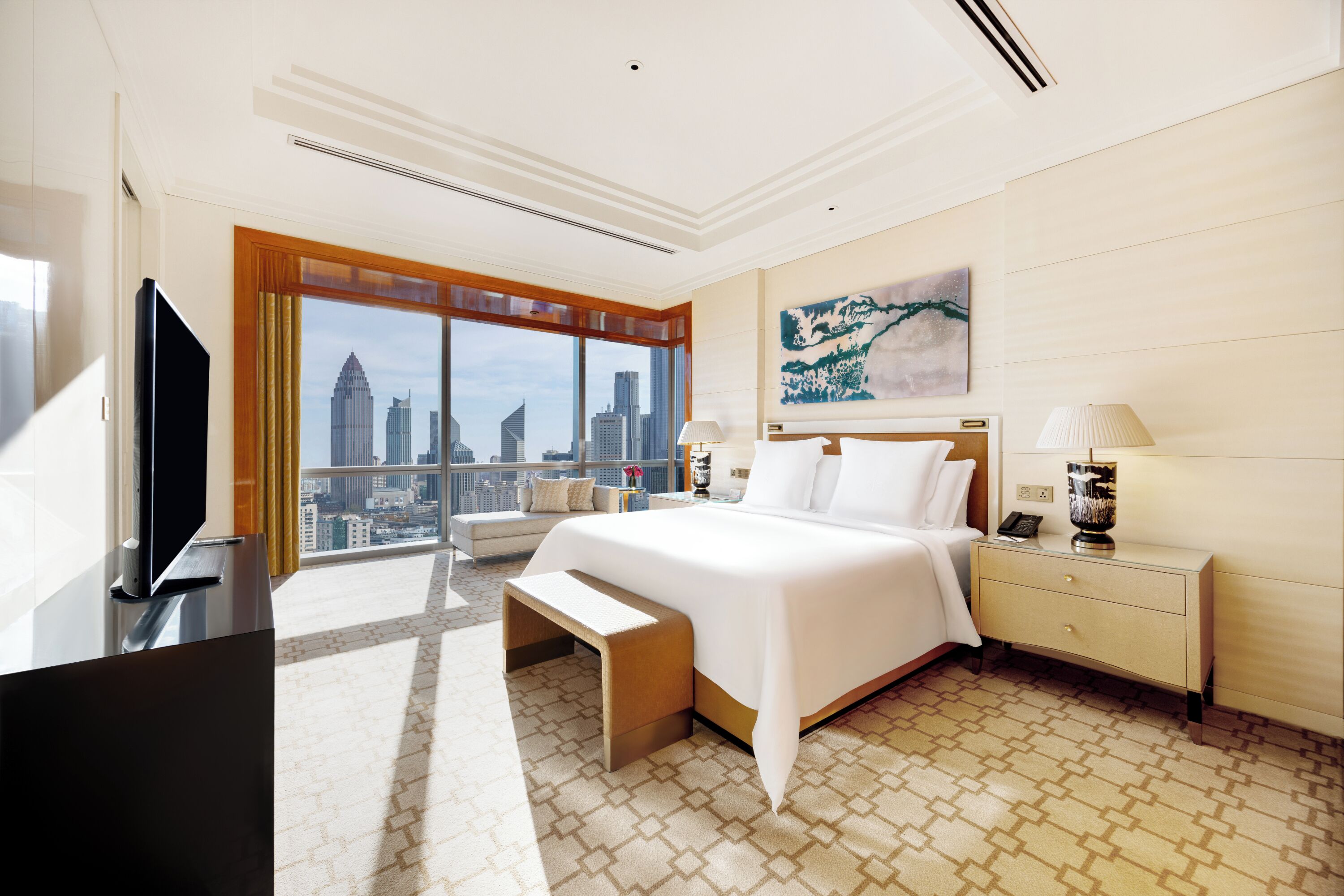 A hotel room with a large bed, a TV, a bedside table with a lamp, and a view of the city skyline.