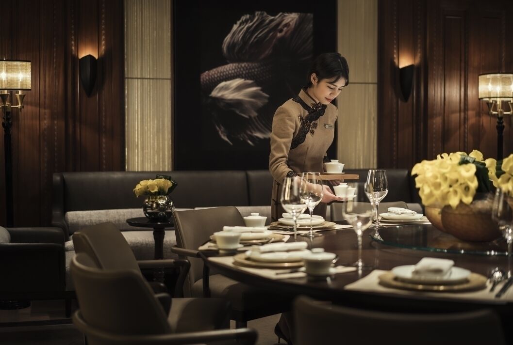 A dining area with a table set for two, a sofa, and a wall-mounted screen displaying a dragon.