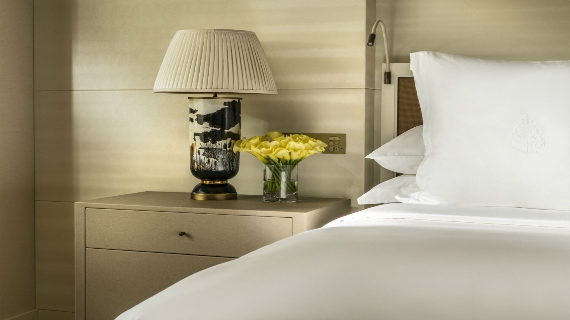 A neatly made bed with white linens, a bedside table with a lamp, and a vase with yellow flowers.