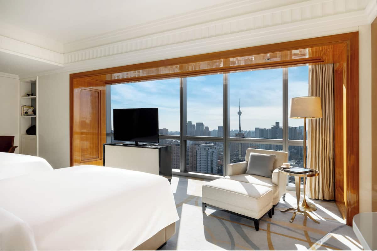 Habitación de hotel con una cama grande, televisión, sofá, una mesita y vistas a un paisaje urbano a través de amplias ventanas.