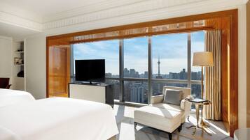 A hotel room with a large bed, a TV, a sofa, a small table, and a view of a cityscape through large windows.