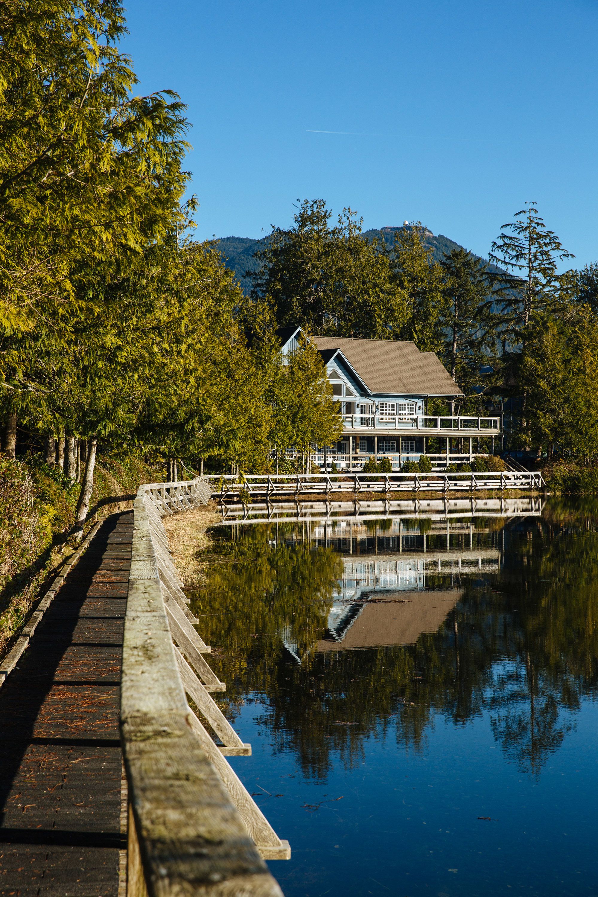 A lakeside wooden pier leading to a house with a balcony, surrounded by trees and a mountain in the background.